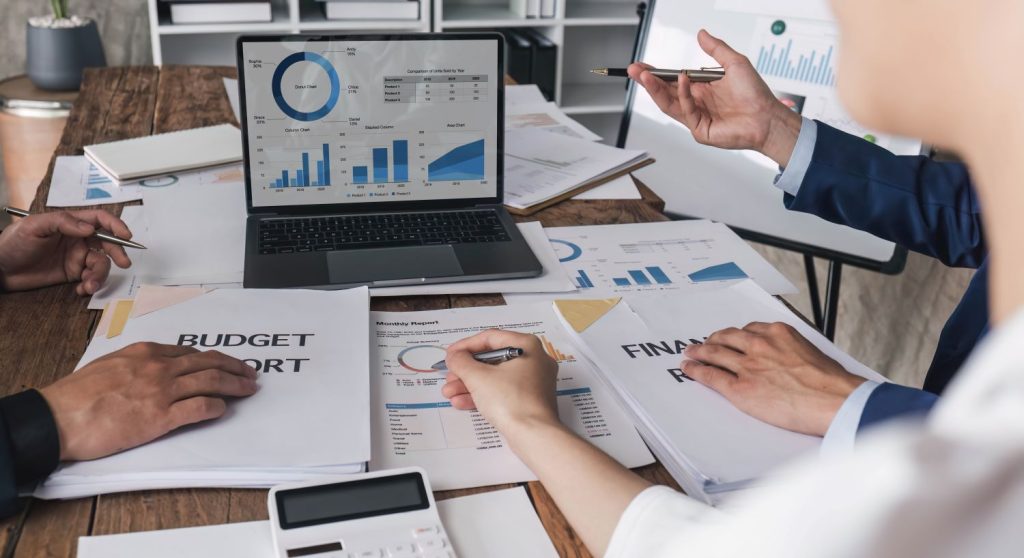 Team meeting around a wooden desk with a laptop showing analytics, scattered budget and financial reports, calculator, and pens — illustrating MSA Asia’s accounting and tax services